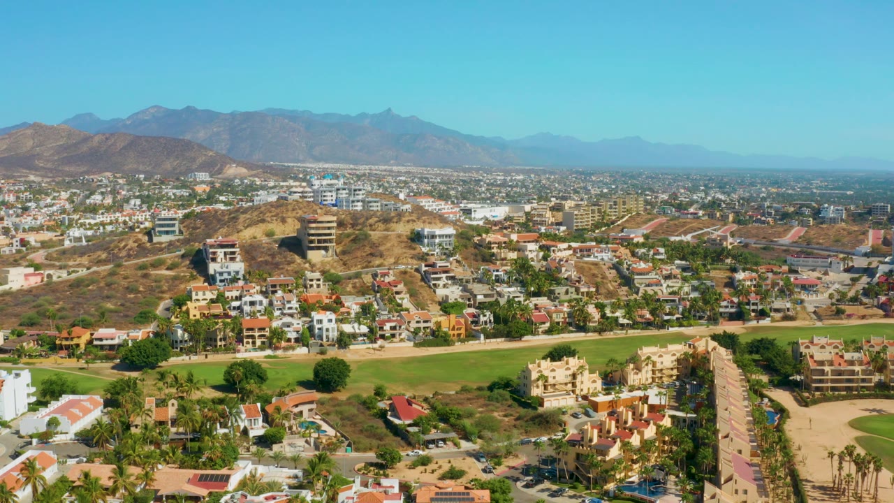 Town aerial with hillside homes, green strip, palms, and hazy mountains.