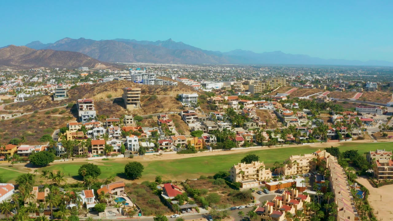 Early frame of San Jose Del Cabo from above, showing a broad green lawn in front of modern homes, dry hills, low-rise town blocks, distant mountains, and no visible people.