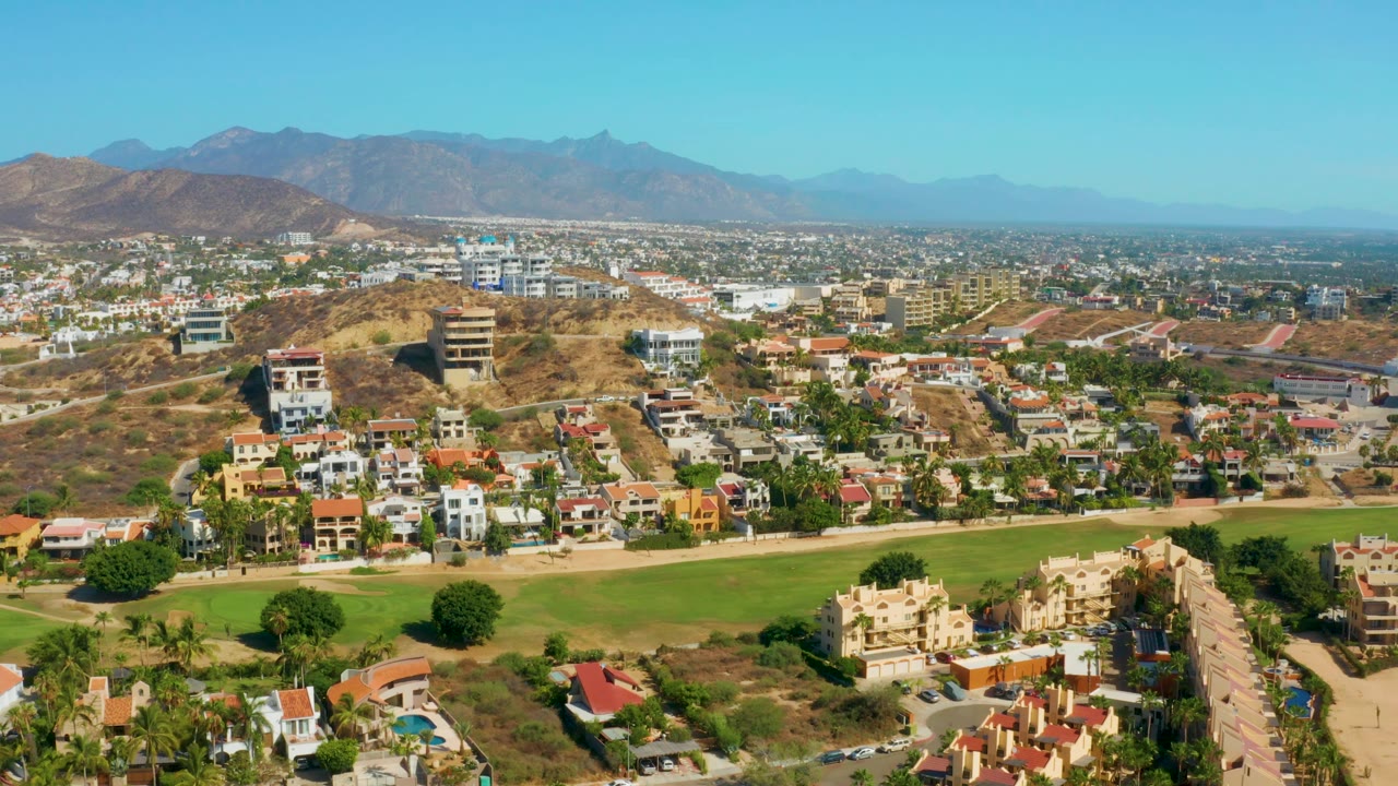 Midwide San Jose Del Cabo view showing the green fairway-like strip across the lower frame, clusters of homes, dry hills, low city blocks, distant mountains, and no visible people.