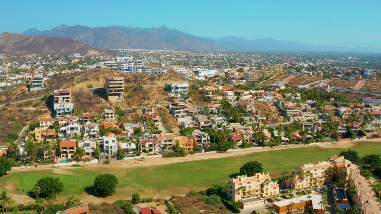 Opening aerial frame over San Jose del Cabo with green lawn in the foreground, hillside houses and streets in the middle distance, brown mountains beyond, and no visible people.