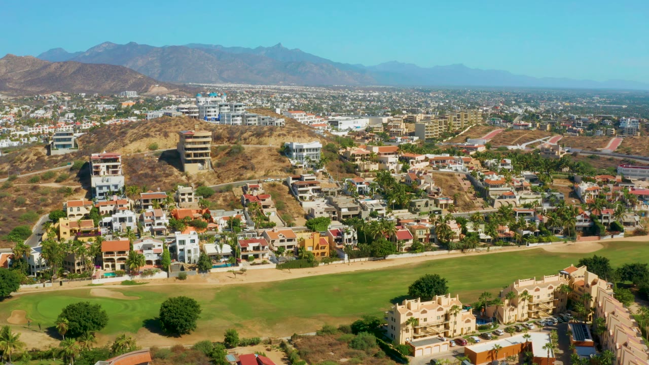San Jose del Cabo aerial view with a broad green foreground, clustered houses, low-rise city buildings, dry hills, distant mountains, and no visible people.