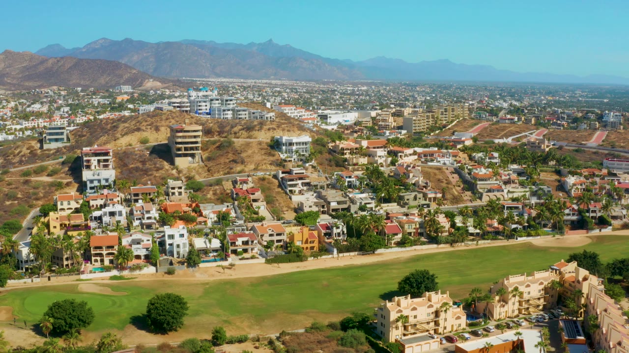 A later aerial frame of San Jose del Cabo shows houses along the lower edge, a large green lawn, denser city buildings across the center, mountains on the horizon, and no visible people.