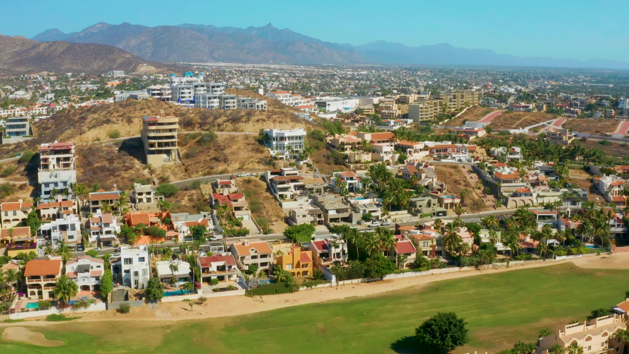 Aerial frame from San Jose del Cabo showing rows of homes and roads below, a dry hill with mid-rise buildings, and blue mountains across the horizon, with no visible people.