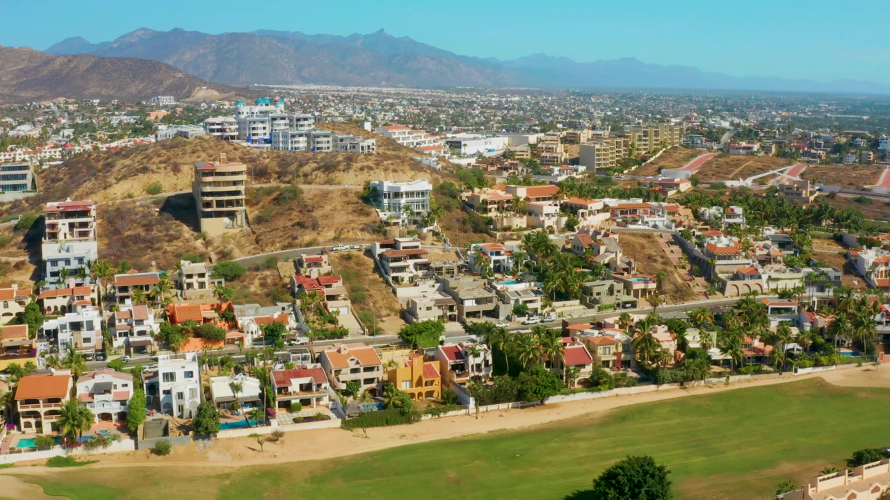 Aerial view of San Jose del Cabo with palm-lined residential blocks in the foreground, tan hillside terrain in the middle distance, and layered mountains under bright blue sky, with no visible people.