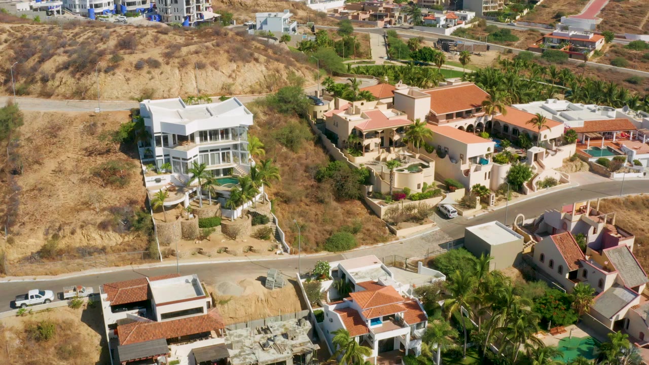 Aerial frame over San Jose del Cabo showing white and terracotta-roofed homes beside a dry hillside road, with palms and no visible people.
