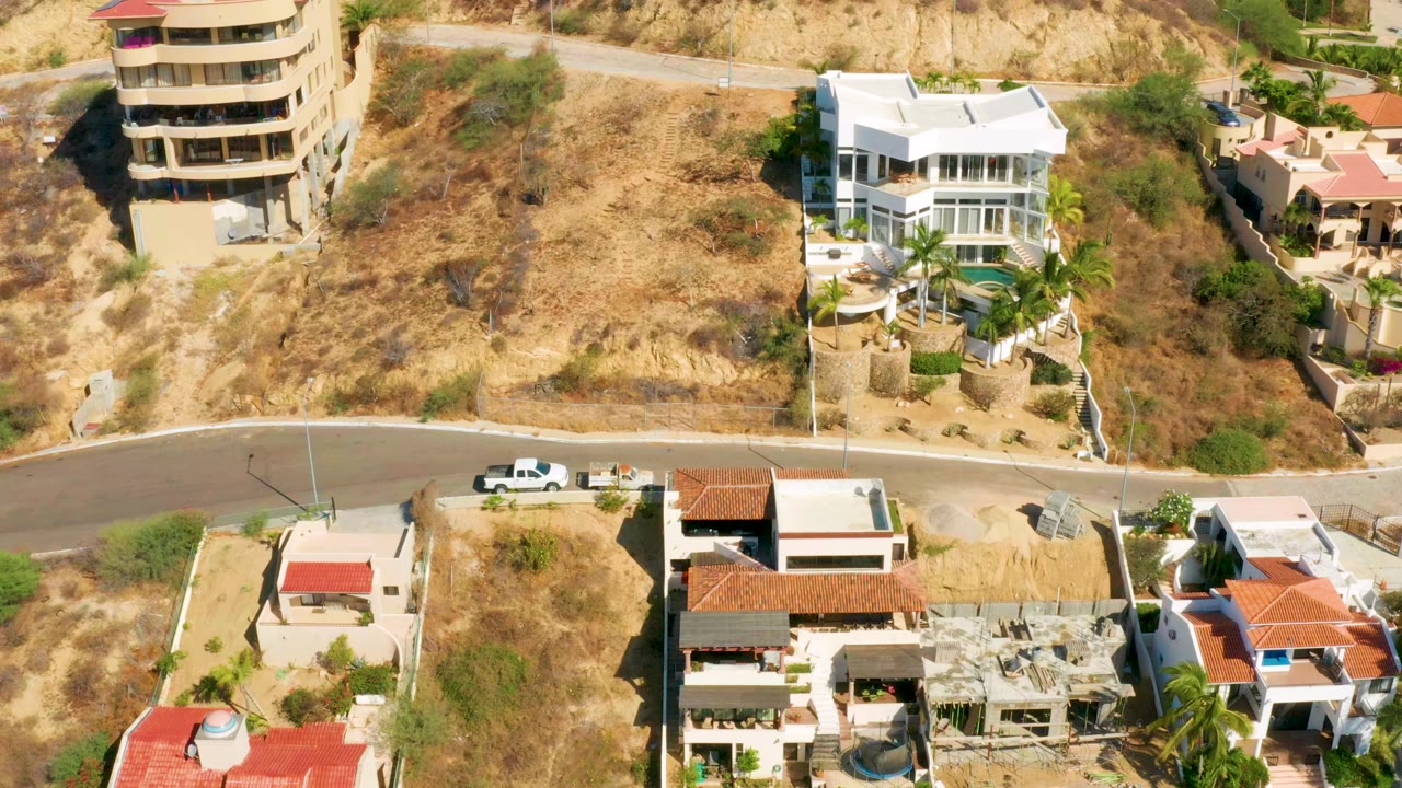 High aerial frame over San Jose del Cabo hillside homes, a curved road, dry lots, white villas, red tile roofs, parked vehicles, and no visible people.