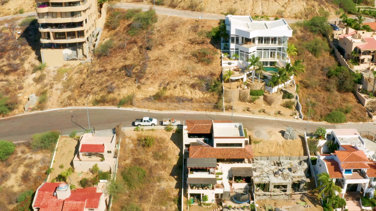 San Jose del Cabo aerial view with red roof homes in the lower frame, a white multi-story house near the hillside, a winding road, dry scrub, and no visible people.