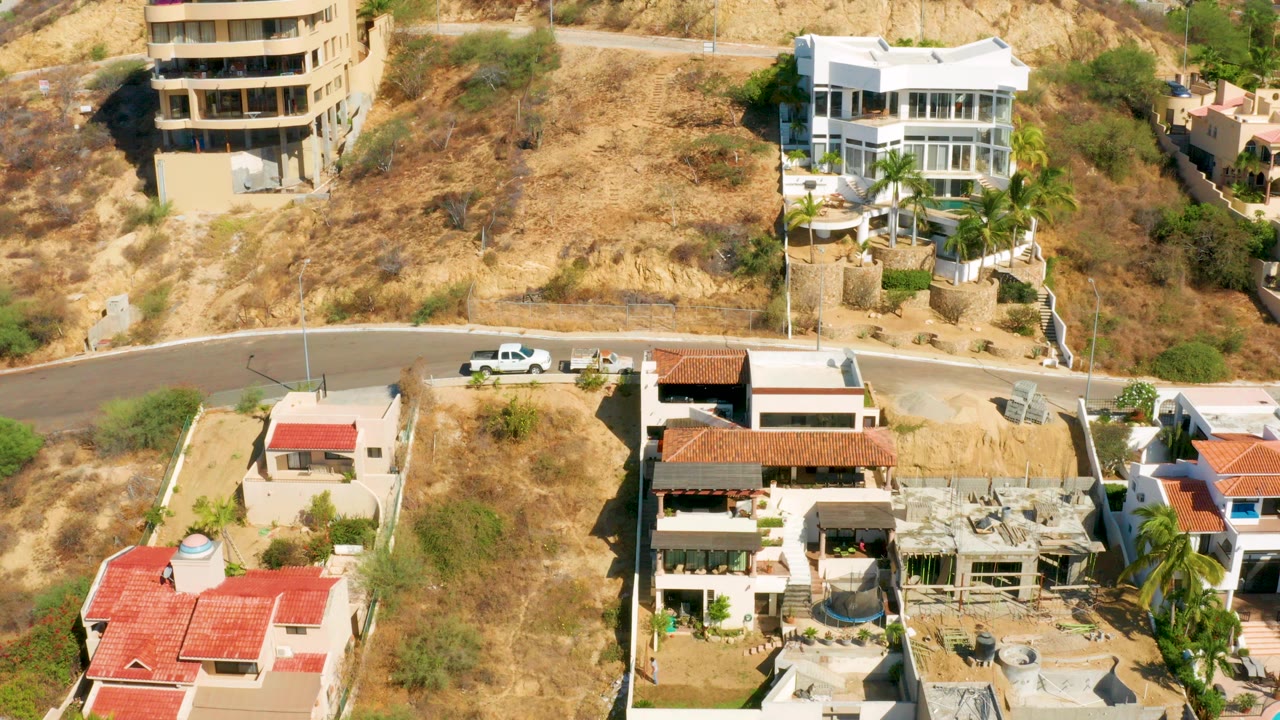 Aerial frame over San Jose Del Cabo hillside homes, dry tan brush, a curved paved road, red tile roofs, and no visible people.