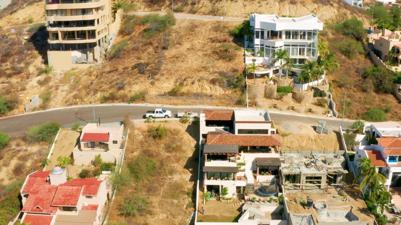 San Jose Del Cabo aerial still with a tan multi-level building, white hillside house, dry vacant slope, paved road, and no visible people.