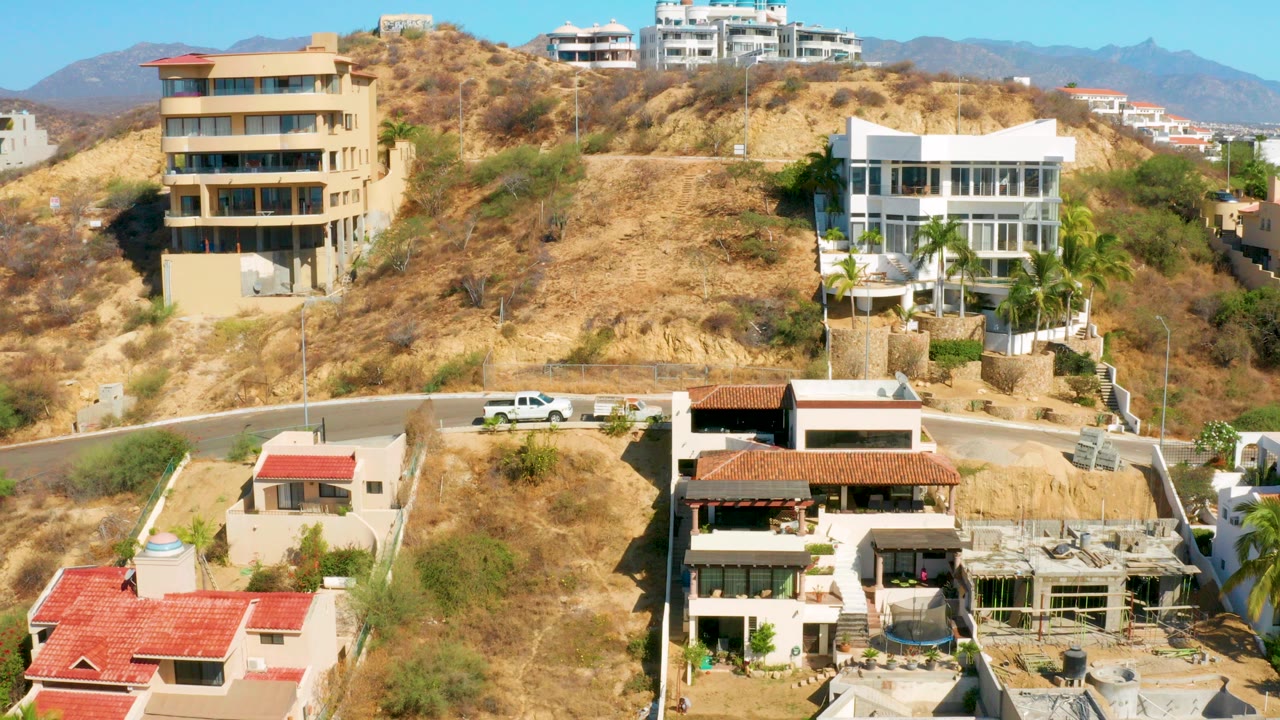 Opening frame of San Jose Del Cabo hillside homes with tan dry slopes, a white truck on the road, distant mountains, and no visible people.