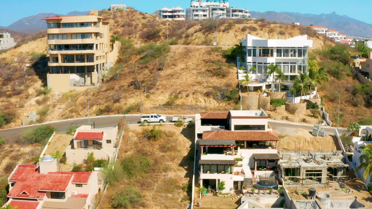 San Jose Del Cabo hillside scene with a tan multi-story building on the left, a white modern house on the right, dry brush, road, and no visible people.