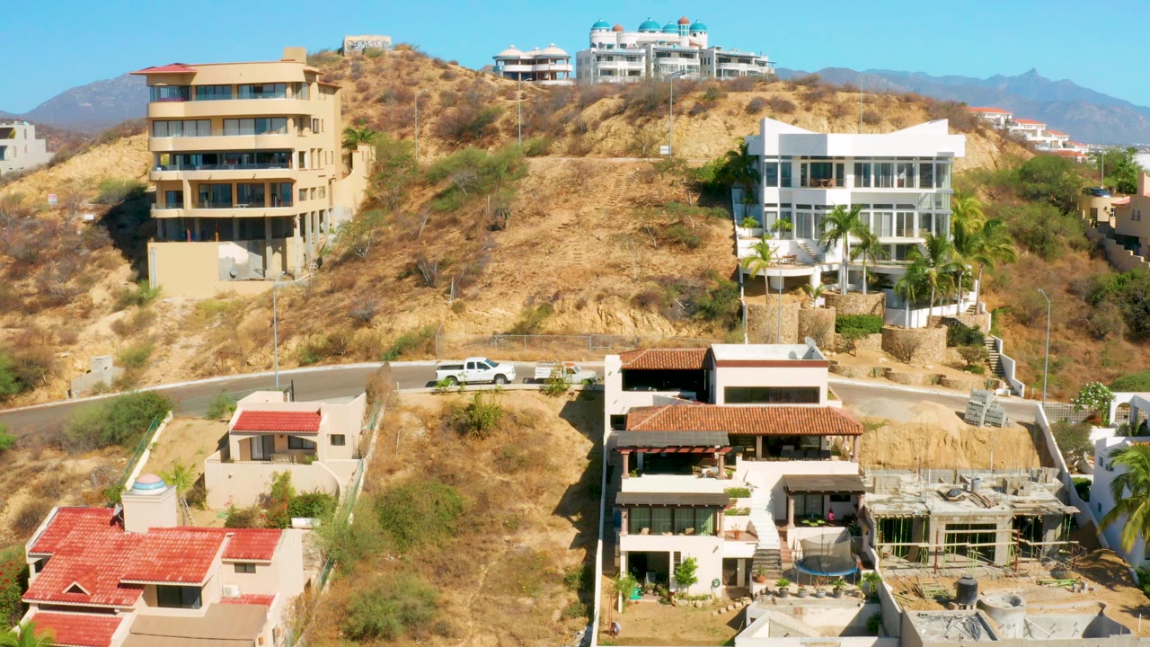 San Jose Del Cabo residential slope with foreground red roofs, hillside homes, a white truck near the road, distant mountains, and no visible people.