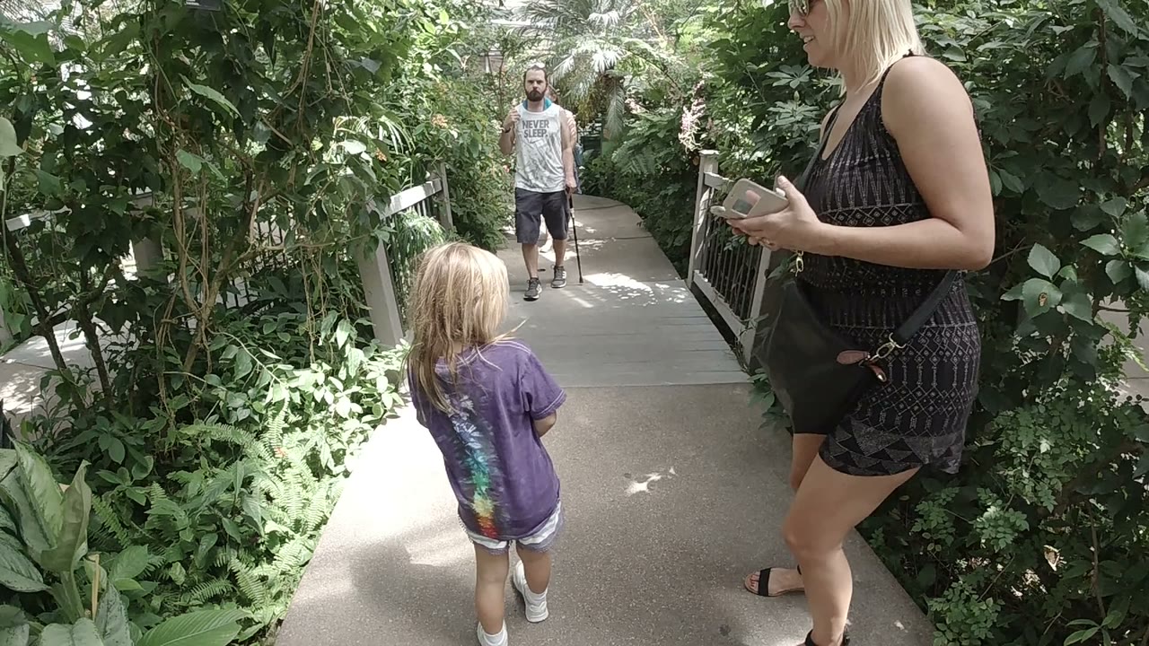 In Denver, Colorado, a blonde child in a purple shirt walks ahead on a narrow path while a woman in a black dress stands cropped at right with a phone in her hands.