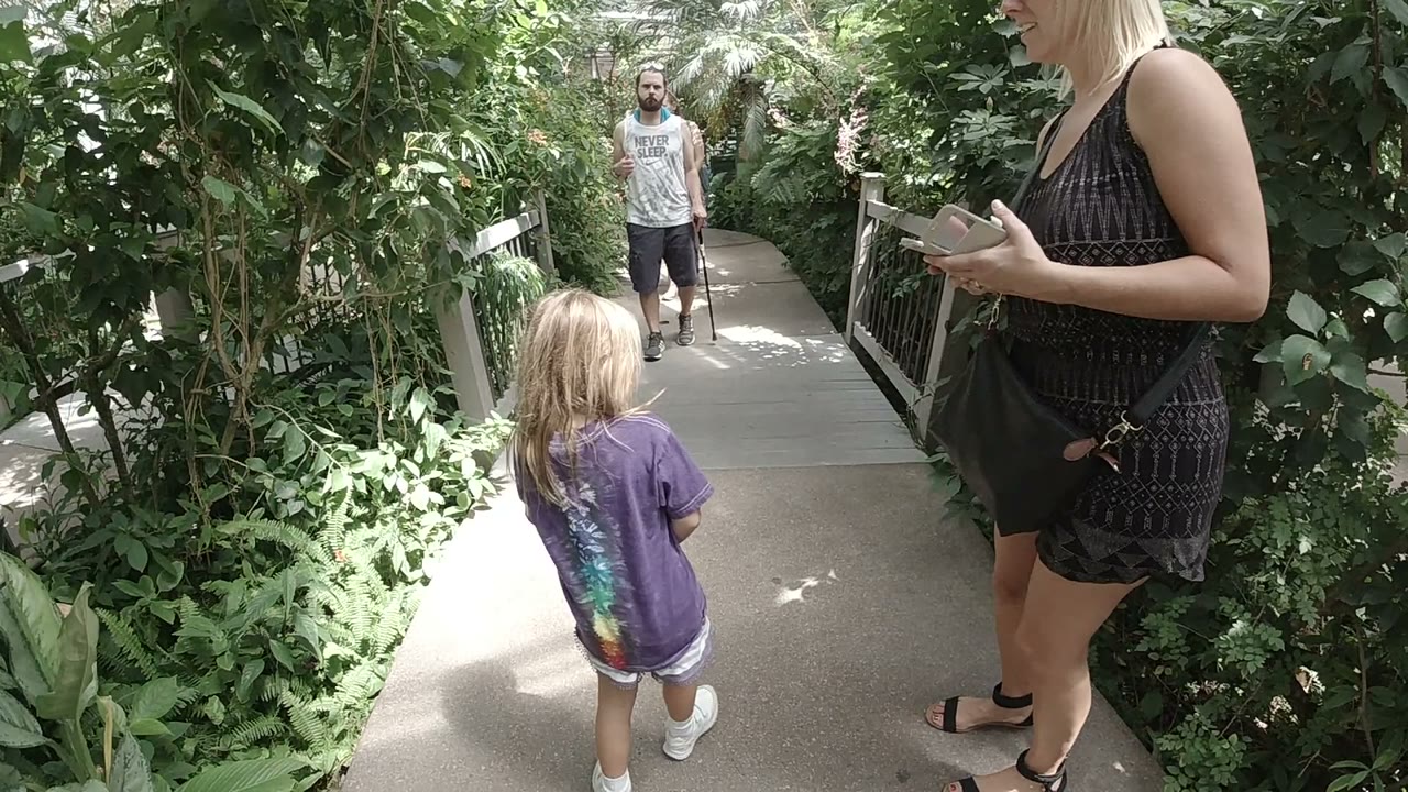 The Denver walkway view holds on the child from behind as a bearded man in a gray sleeveless shirt approaches from the shaded path ahead.