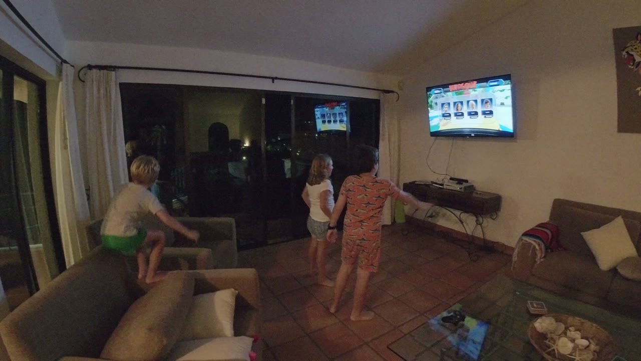 In the Los Cabos interior, one child climbs on a sofa while two others stand near the center of the room facing the bright television.