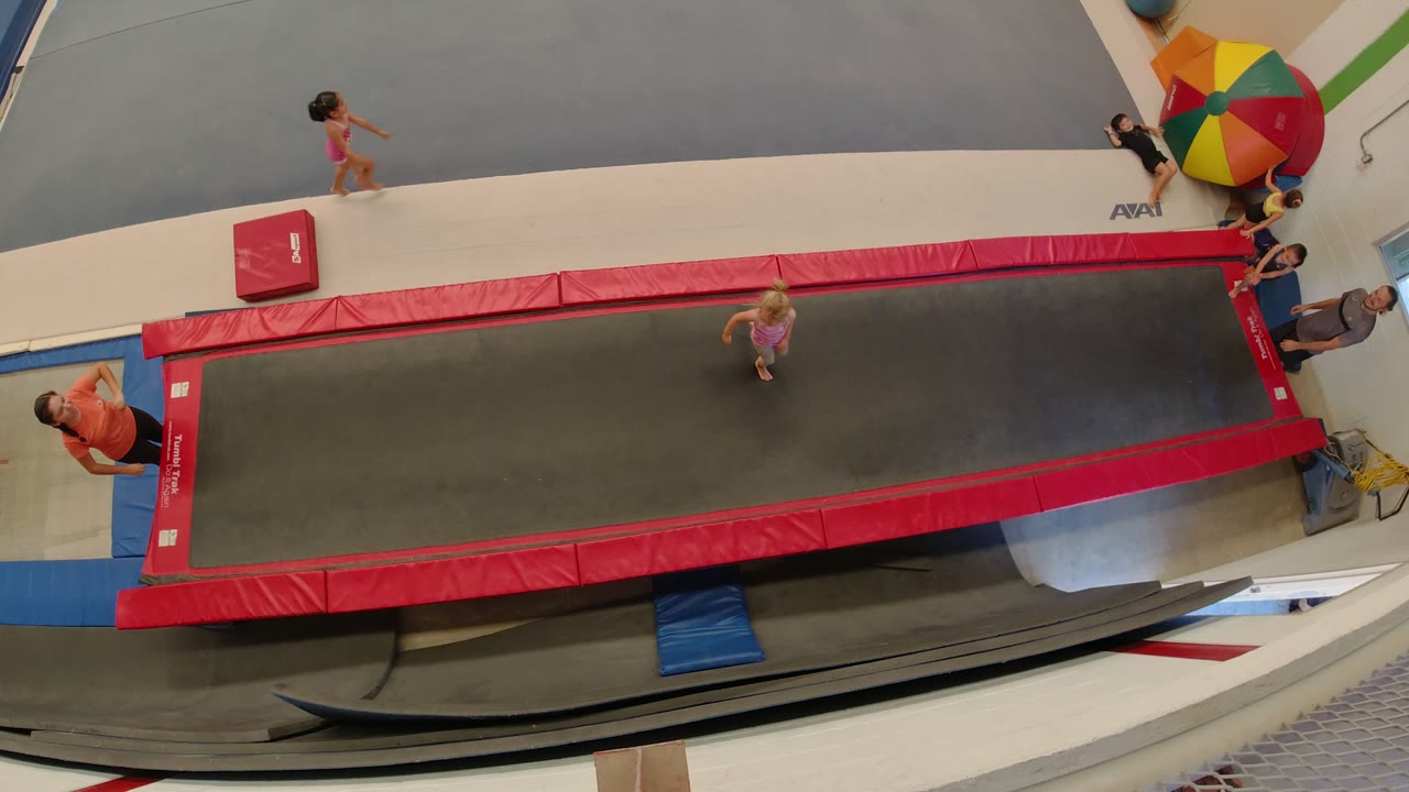High angled Los Cabos frame showing a child on the trampoline surface, another child walking nearby, and an adult coach positioned along the red padded edge.