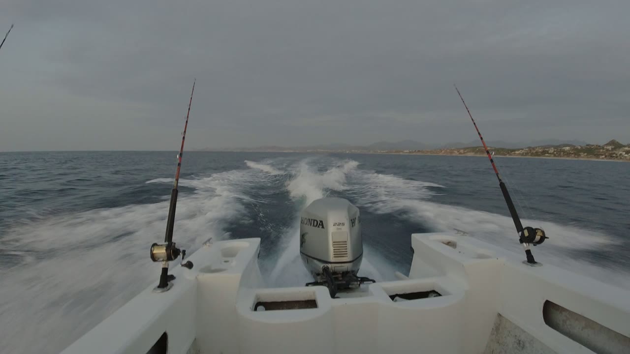 At 0:00, two fishing rods frame a white outboard motor as the boat leaves a foamy wake off Los Cabos, Mexico, with no visible people.
