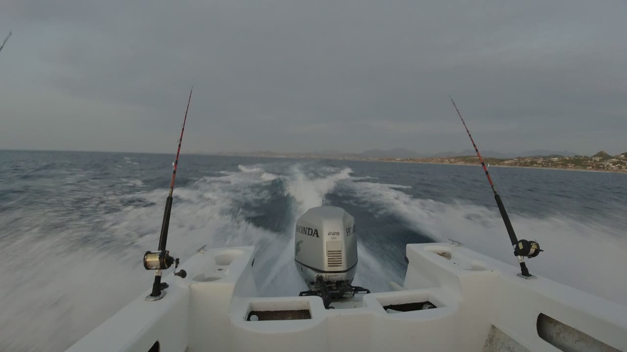 At 0:01, the stern view holds on the paired fishing rods and centered outboard motor while the wake spreads across the water near Los Cabos, Mexico, with no visible people.