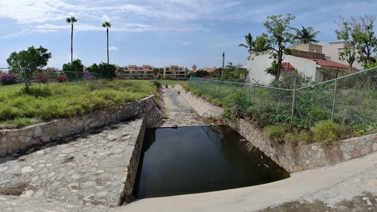 Opening frame of a dry aqueduct in Los Cabos, Mexico, with a shallow dark pool in the foreground, grasses and chain-link fencing beside the channel, distant buildings, and no visible people.