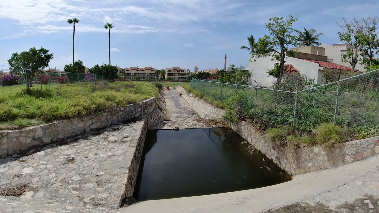 The aqueduct remains centered in Los Cabos, Mexico, with the stone-lined path leading toward distant buildings, palms, scrubby grass, a fence line, and no visible people.