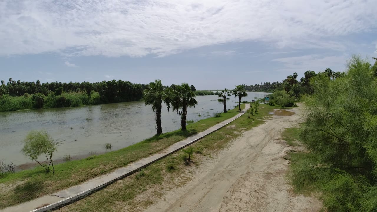 Elevated view in Los Cabos of a canal or intercoastal waterway beside a pale dirt road, sidewalk, palm trees, and dense green brush, with no visible people.