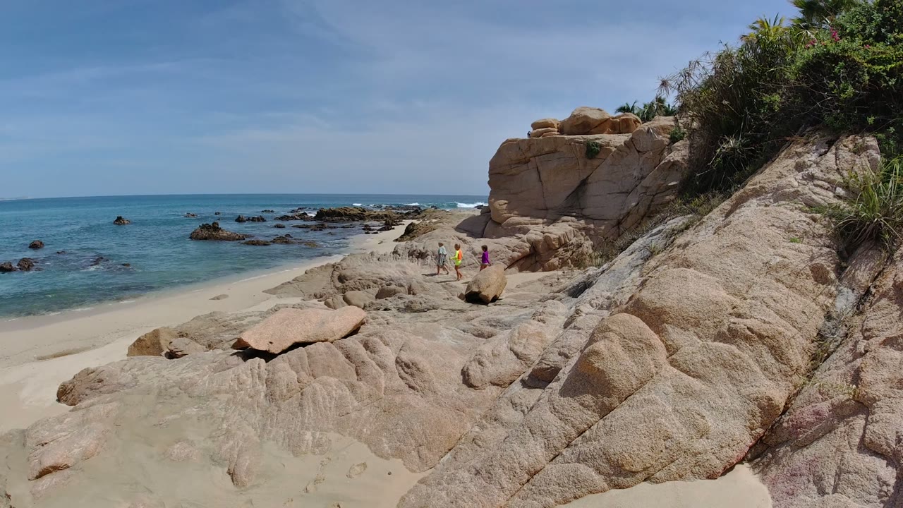 Los Cabos, Mexico shoreline with tan foreground rocks, blue ocean to the left, a scrubby rock slope to the right, and two distant people walking near the sand.