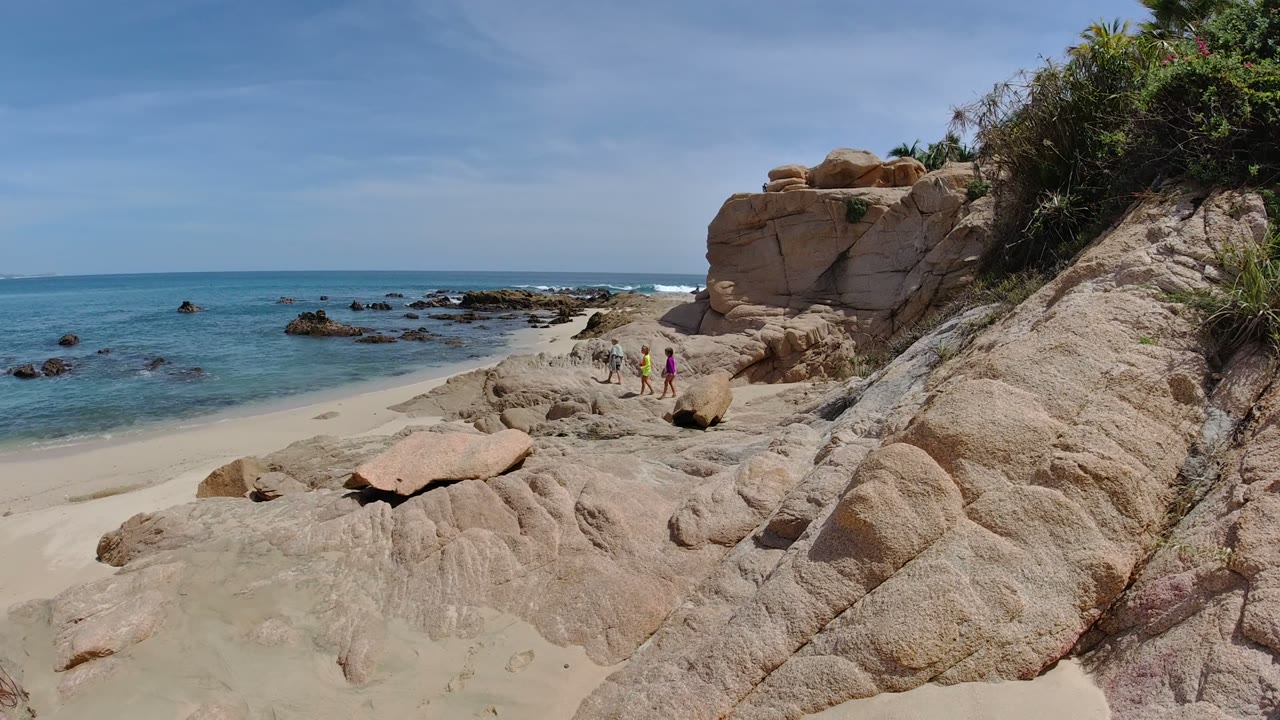 Los Cabos, Mexico beach view with rounded tan rocks across the foreground, low waves near scattered offshore rocks, and two small people slightly farther along the shoreline.