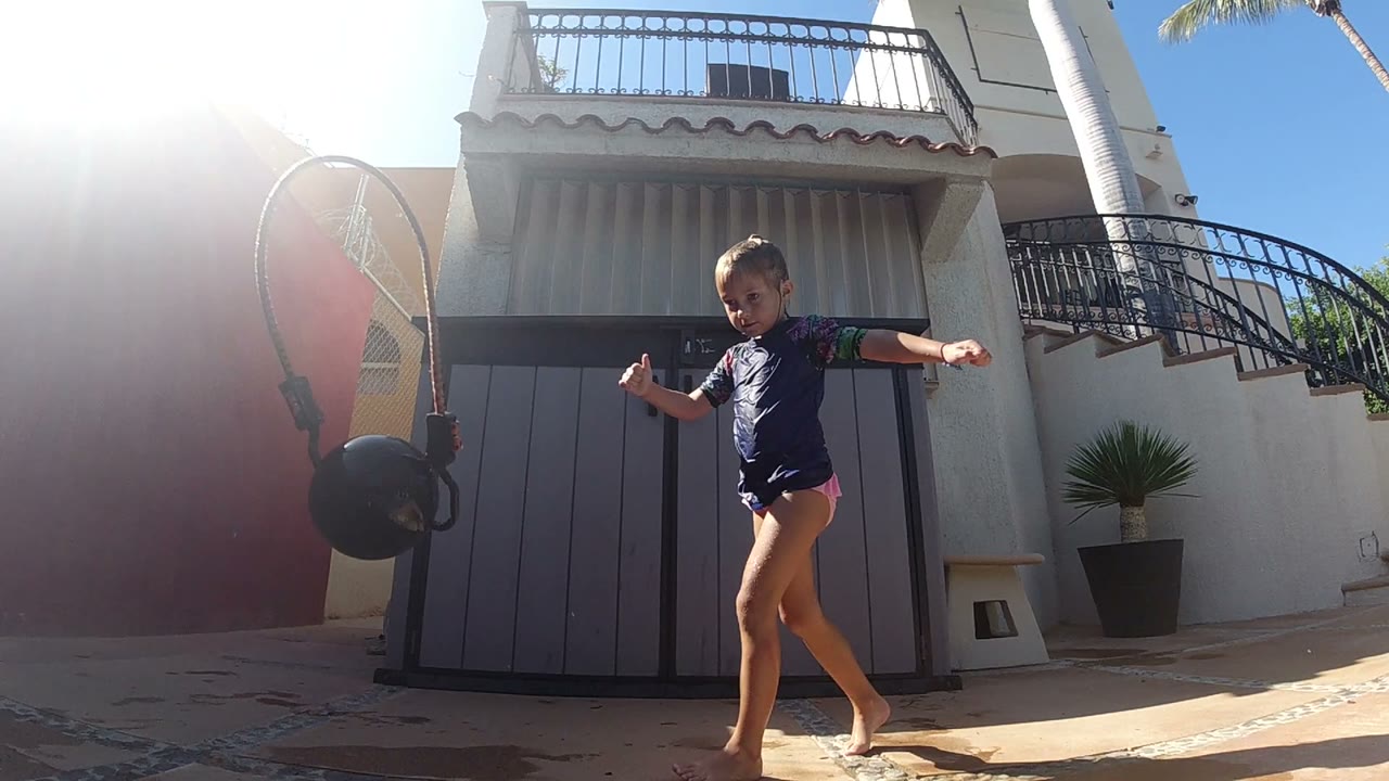 The girl steps forward in Los Cabos with the black ball suspended near the left edge, strong backlight washing the red wall and gray doors behind her.