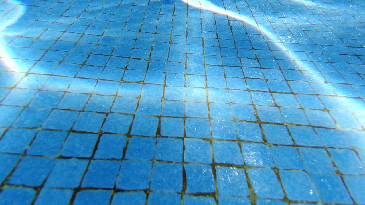 Blue square pool tiles fill the Los Cabos underwater frame as diagonal white caustic lines cross the lower left, with no visible people.