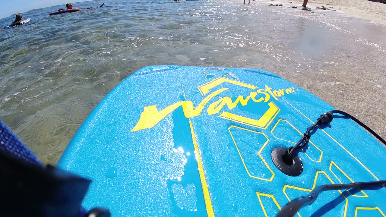 A blue board with yellow graphics fills the foreground in Los Cabos, Mexico, with swimmers in shallow water and beachgoers along the bright sandy shoreline.