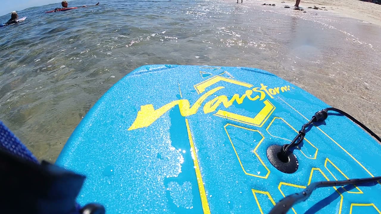 The blue board remains close to the lens in Los Cabos, Mexico, as rippled shallow water reflects sunlight and small figures stand or swim near the shoreline.