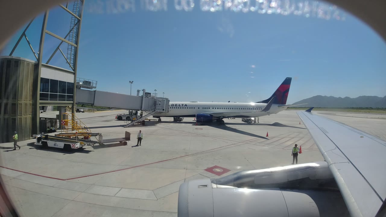 At 0:01 in Los Cabos, Mexico, the airplane wing points toward a Delta jet connected to a jet bridge, with a baggage or service cart near the terminal and workers spaced across the ramp.