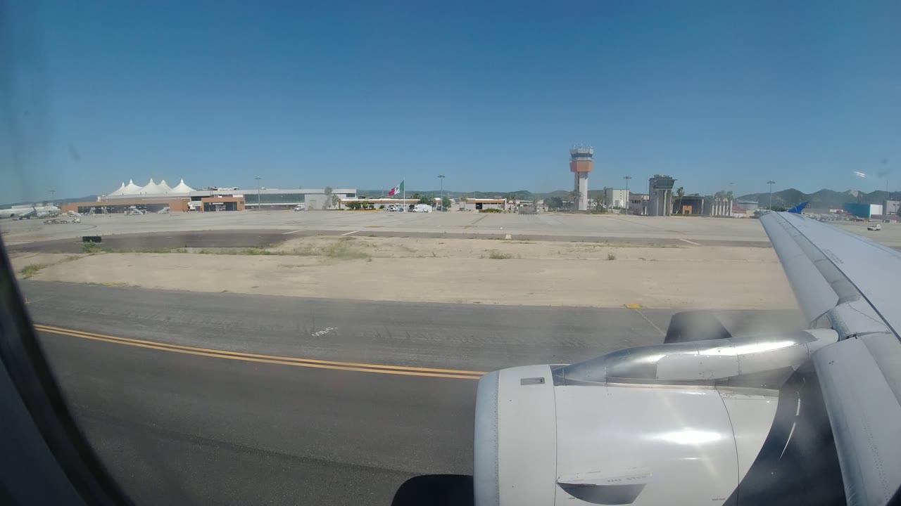 Opening frame from a passenger window in Los Cabos, Mexico, showing the aircraft wing and engine over runway pavement, sandy ground, terminal buildings, a control tower, and no visible people.