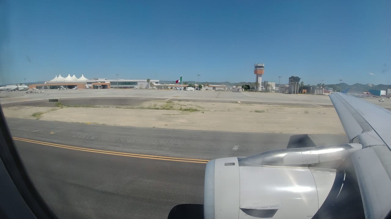 A second passenger-window view in Los Cabos, Mexico, keeps the wing and engine in the foreground as terminal roofs, the control tower, distant planes, mountains, and no visible people remain in view.