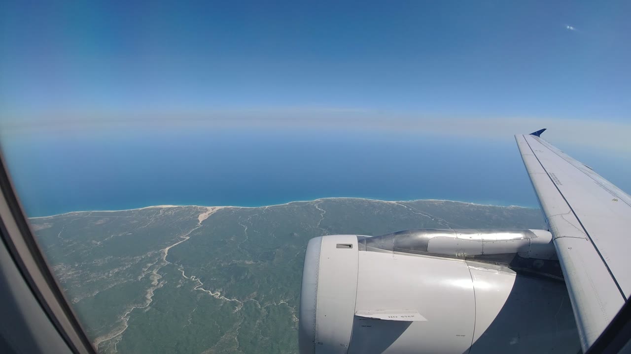 Airplane wing and engine over coastline and blue ocean.
