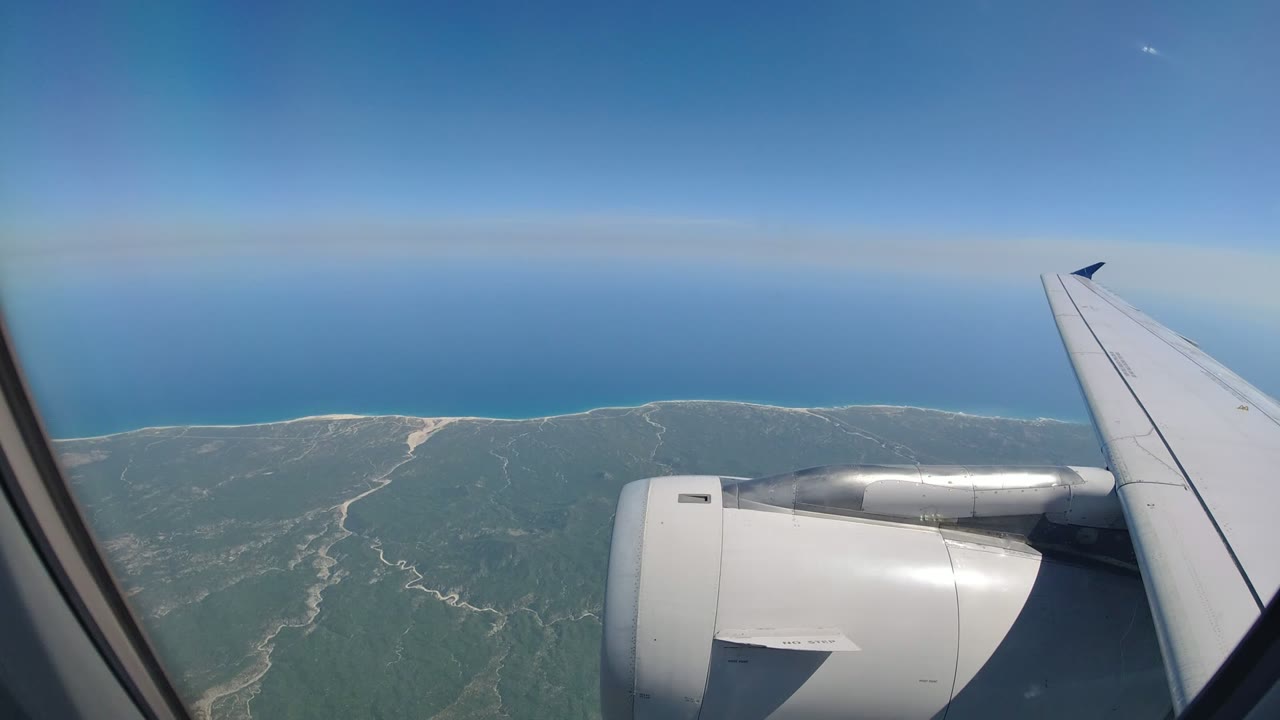 Los Cabos coast seen from a passenger window, with the engine nacelle centered low, the wing angled upward, ocean on the horizon, and no visible people.