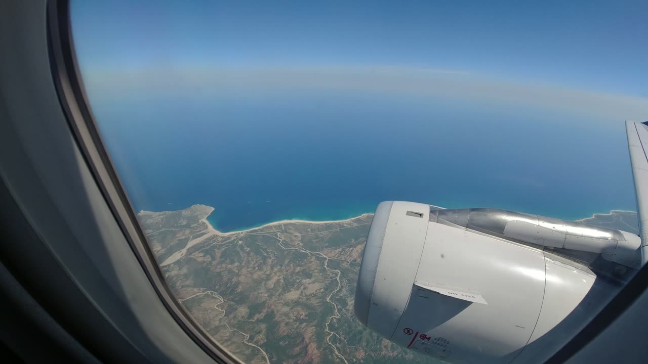 Passenger-window view over Los Cabos, Mexico, with the airplane engine at right, rugged coastline below, blue ocean beyond, and no visible people.
