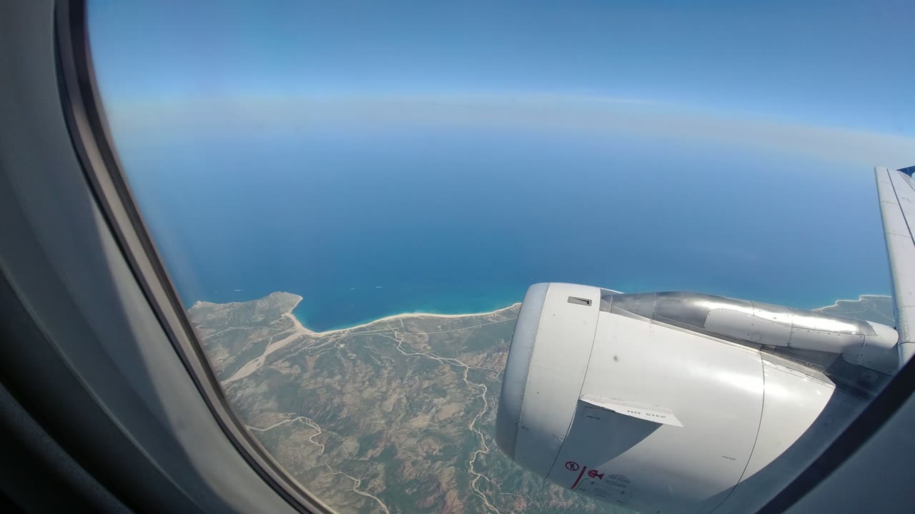 The aircraft engine fills the right side of the window view while the Los Cabos coastline curves below under a clear blue sky, with no visible people.
