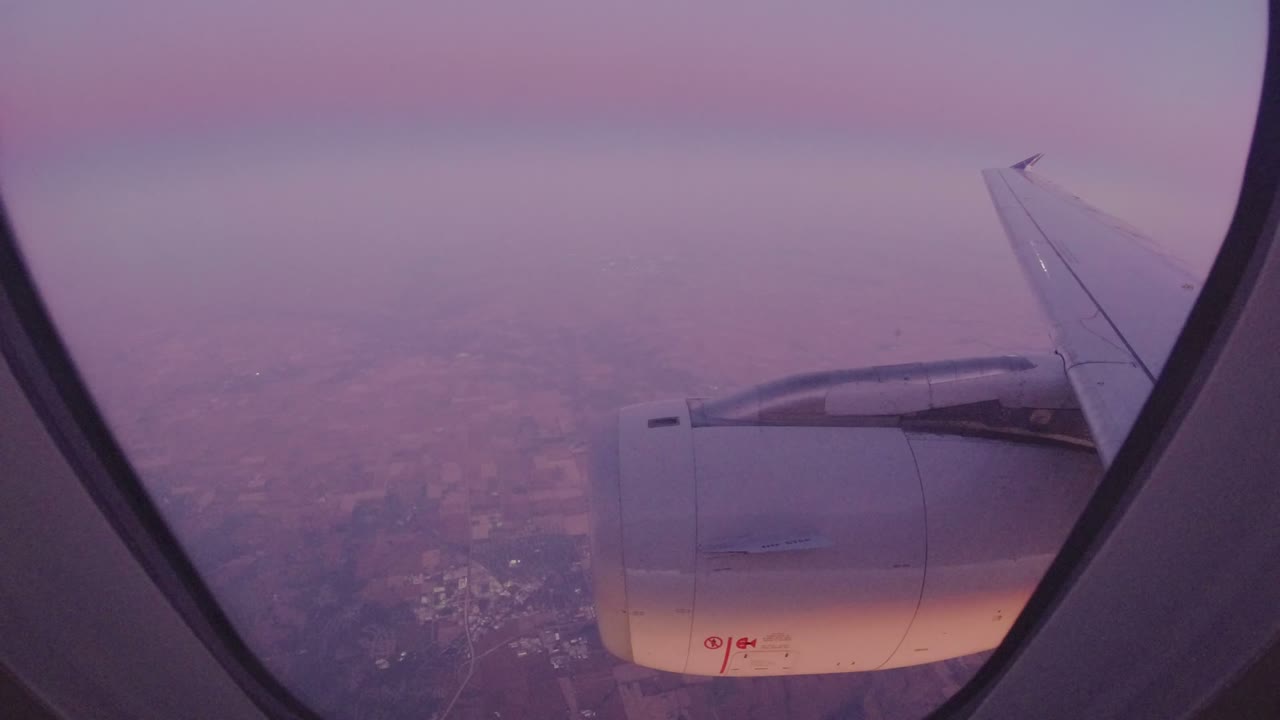 Airplane wing and engine seen through a passenger window above hazy land.
