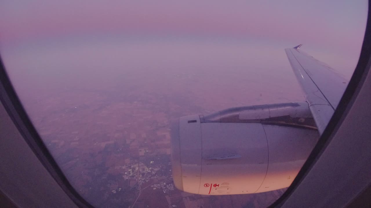 One second into the Los Cabos, Mexico clip, the airplane engine remains centered low in the window, the wing points toward the upper right, and the hazy landscape sits beneath a purple cast with no visible people.