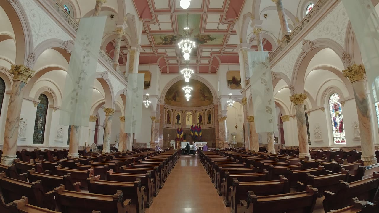 Wide view inside a Los Cabos church shows dark wooden pews leading toward an altar, with arched columns, chandeliers, stained glass, and a small distant group of people at the front.