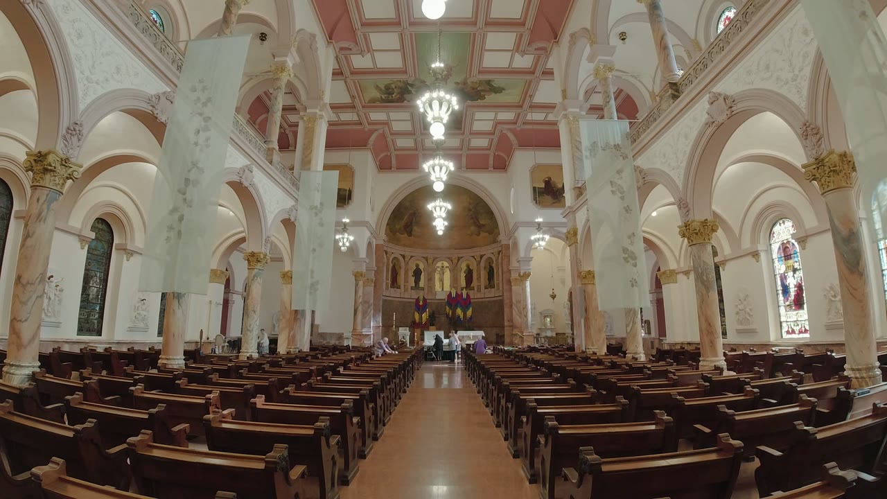 A centered Los Cabos church interior frame shows the central aisle, rows of dark pews, pale columns, hanging lights, stained glass, and people gathered near the altar.