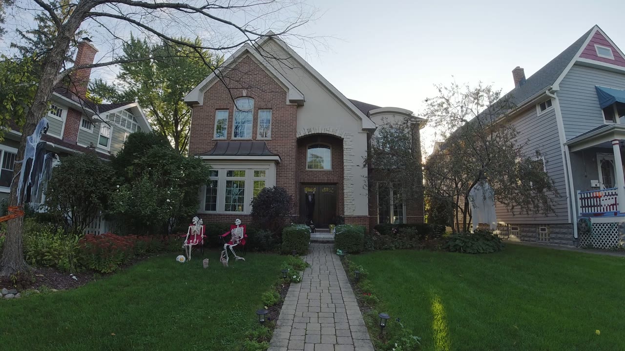 Wide view of a Los Cabos front yard with green grass, a paved walkway, a large tree, a brick house entrance, neighboring homes, and two skeleton-like yard decorations near the lawn.