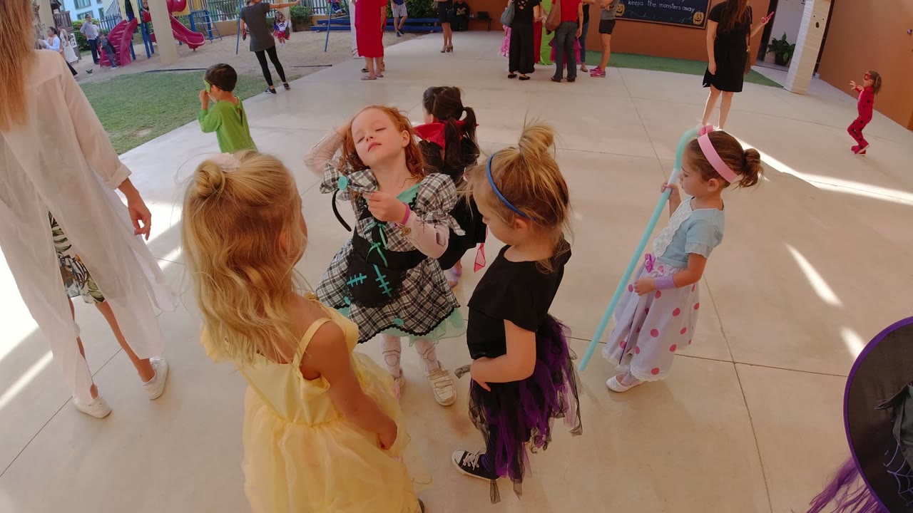 Young children in costumes gather on a sunny paved play area.