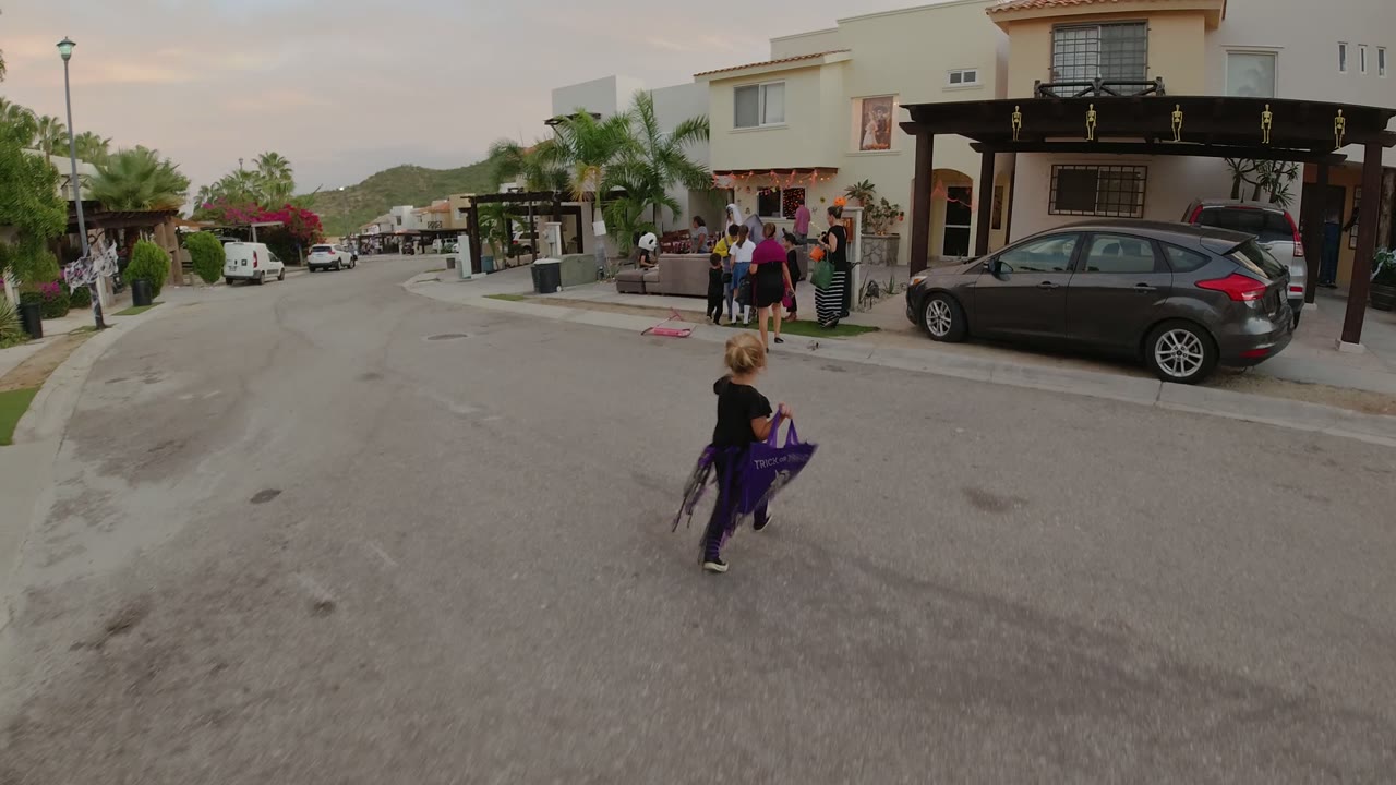 A child carrying a purple bag moves farther along the Los Cabos residential street toward adults and children standing near a cream-colored house.