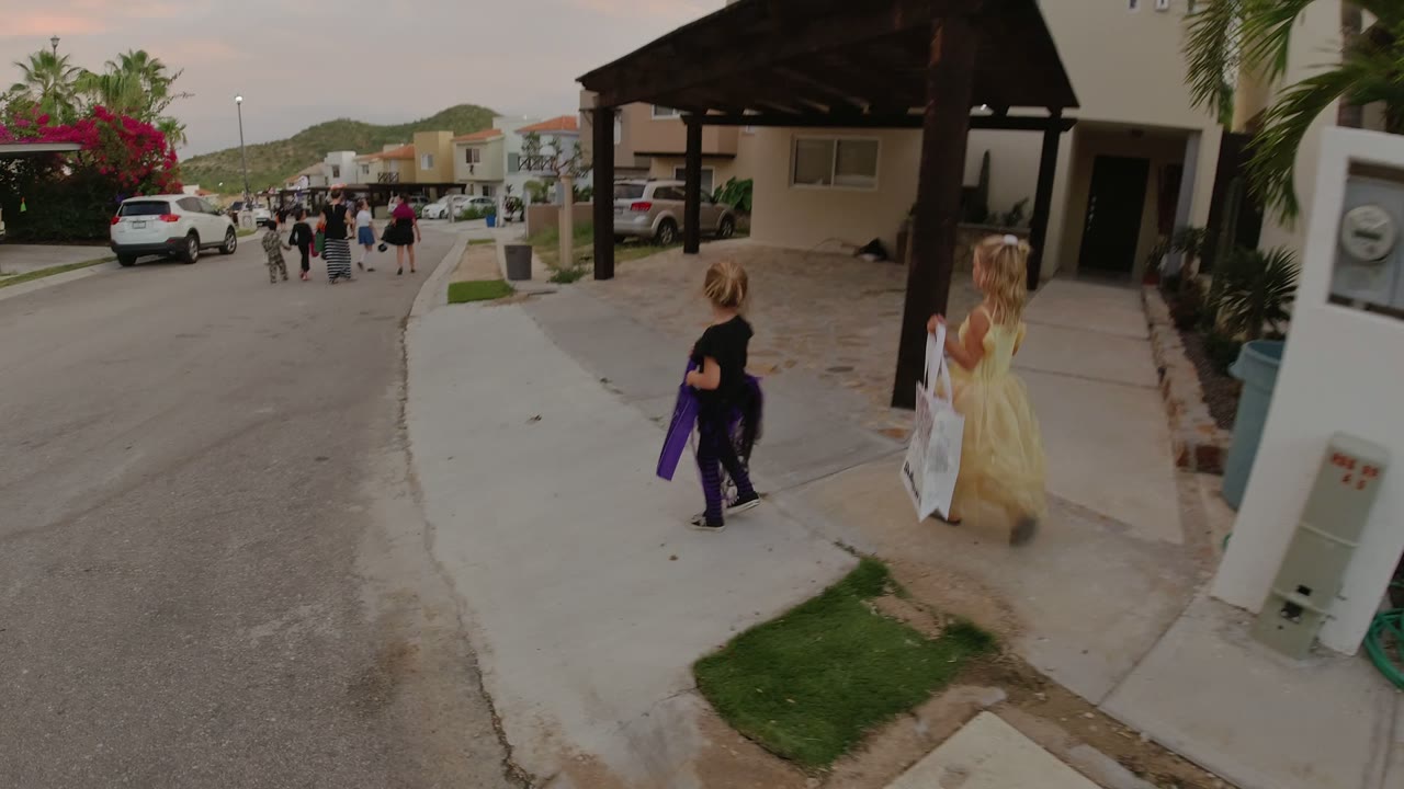 The girls pass a white utility box and hose on a Los Cabos sidewalk while other pedestrians move farther down the street.