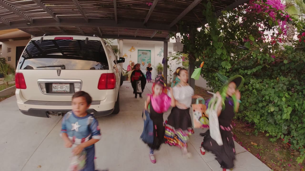 Children in costumes run past a white SUV in a covered driveway.