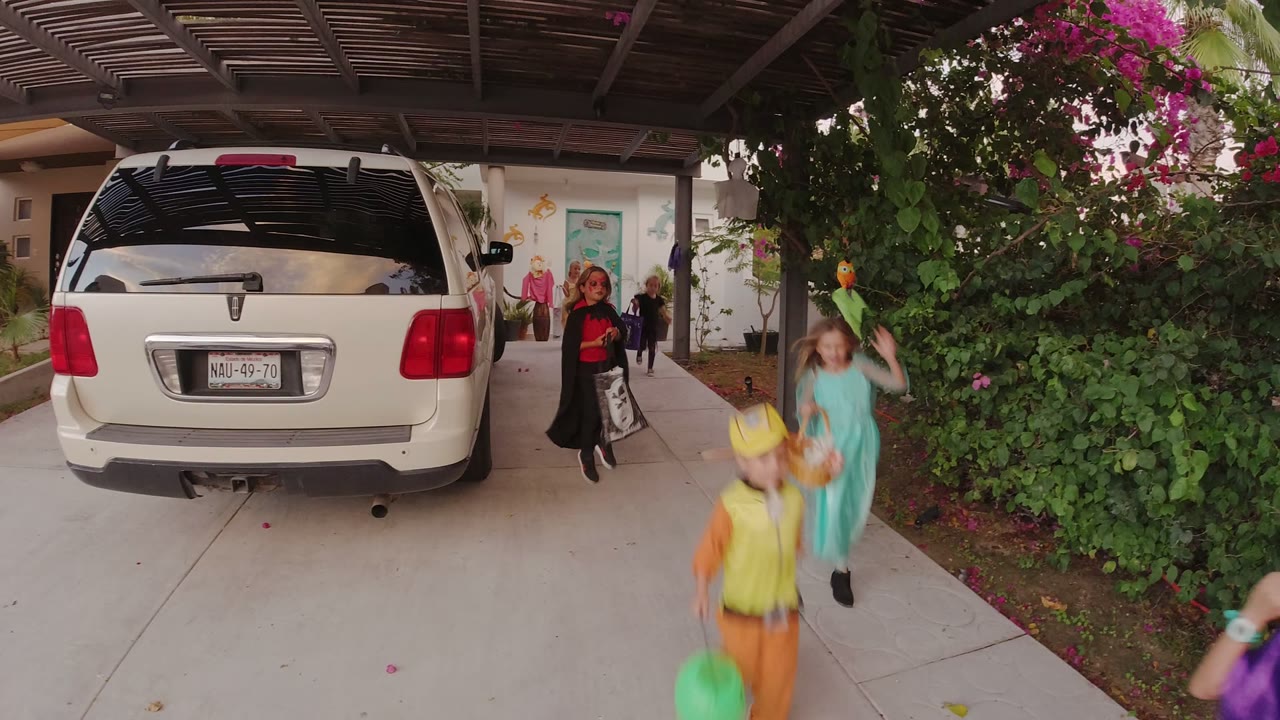 A child in a yellow costume and another child in teal pass the foreground in Los Cabos as adults stand near a turquoise door behind a white SUV.