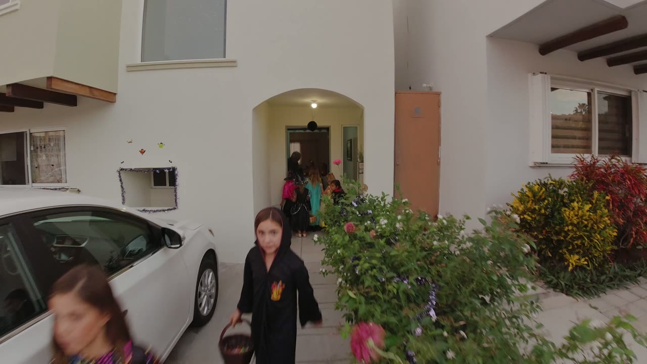Children in Halloween costumes move near a covered residential doorway in Los Cabos, with a white car, garden shrubs, and cream exterior walls visible.