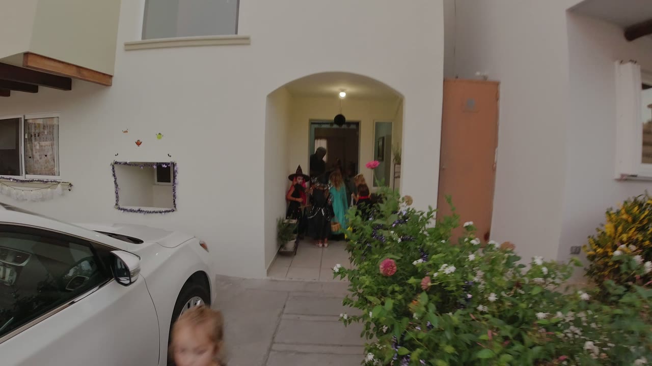 A group of costumed children stands inside the recessed entrance in Los Cabos while a parked white car and flowering shrubs frame the foreground.